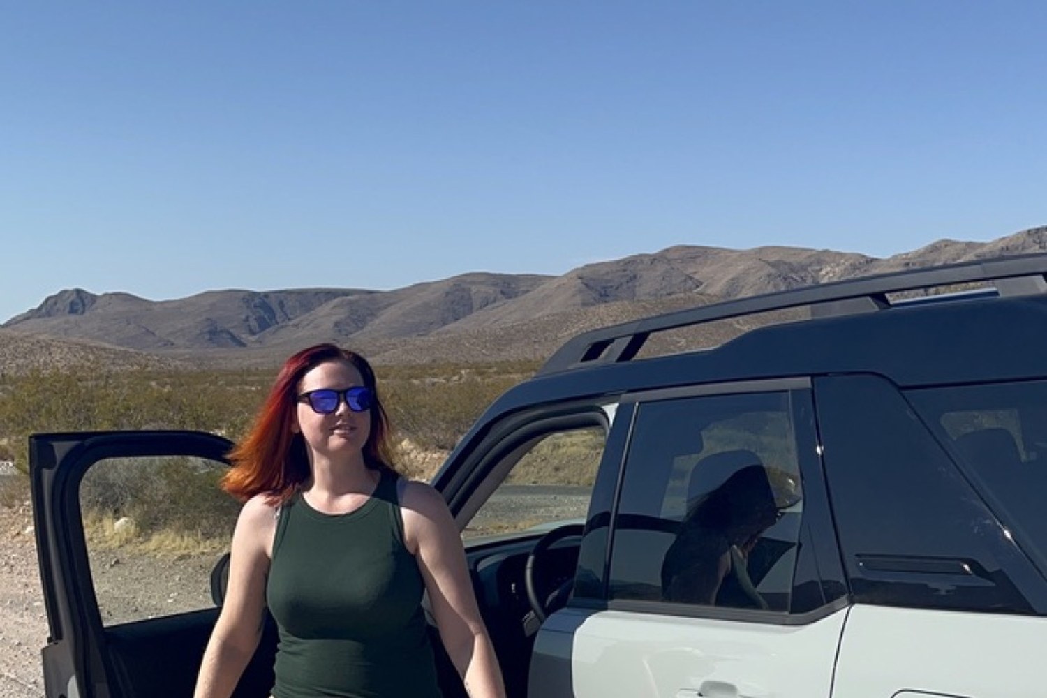 Standing beside Daisy the Bronco with its door open on a desert pullout, mountains behind